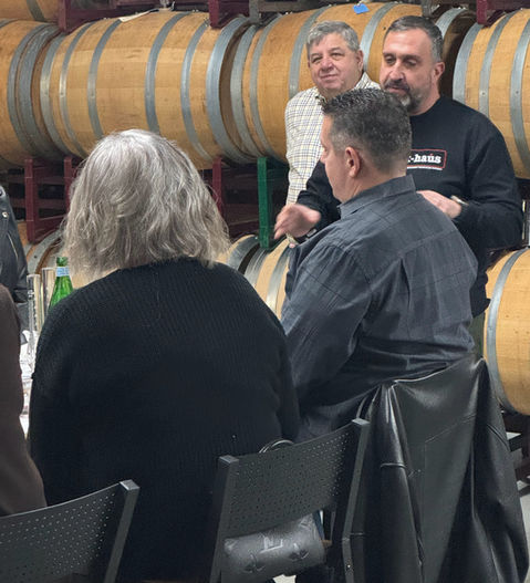 Group of people in front of wine barrels, discussing wine together.