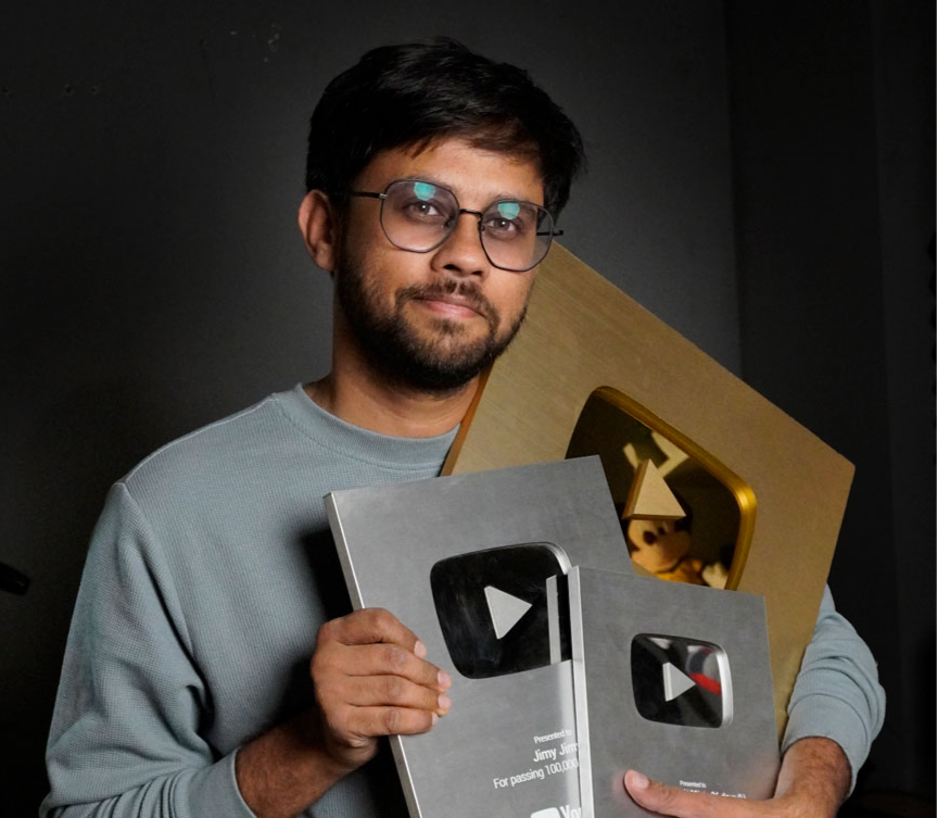 Man, content creator smiling, holding two silver YouTube play buttons and one golden play button against a dark background. He's wearing glasses and a gray sweater. Jaimin Suthar