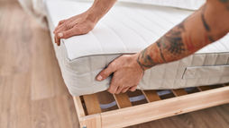 Tattooed arms adjusting a white mattress on a wooden bed frame in a bright room, representing the best Mattress Toppers in memory for Back Pain Relief.