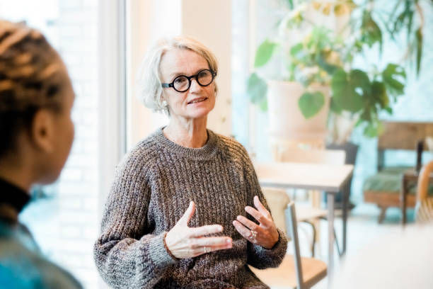 Older woman in glasses and a gray sweater speaking warmly to another person in a bright room with plants, symbolizing connection and guidance in holistic medicine careers.