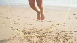 Bare legs and feet mid-jump over a yellow rope on a sandy beach, with the ocean blurred in the background.