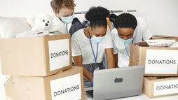Social workers wearing face masks collaborate around a laptop in a donation center with labeled donation boxes and a teddy bear.