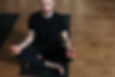 Man in a black shirt sitting cross-legged on a black mat on a wooden floor, practicing zen practice meditation with a calm and focused expression.