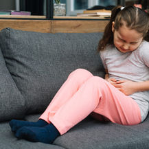 Young girl in pink pants sits on a gray couch, clutching her stomach and grimacing in pain.