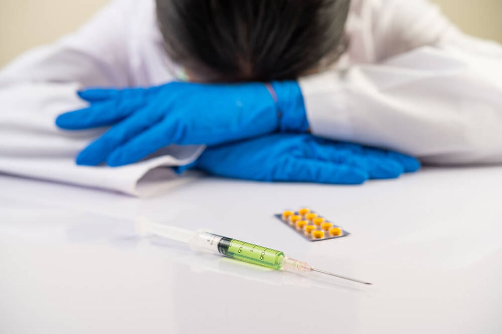 Person in a lab coat with blue gloves rests head on table. Foreground shows a syringe with green liquid and yellow pills. Calm mood.