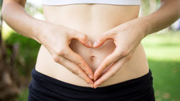 Hands forming a heart shape over a woman’s bare midriff, wearing a white top and black pants, with a blurred green outdoor background, symbolizing collagen and gut health.