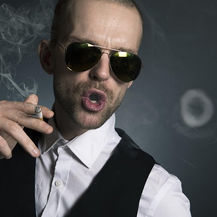 Man in sunglasses blows smoke ring, holding cigarette. Wears white shirt, black vest. Dark background, relaxed and confident mood.