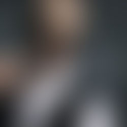 Man in sunglasses blows smoke ring, holding cigarette. Wears white shirt, black vest. Dark background, relaxed and confident mood.