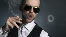 Man in sunglasses blows smoke ring, holding cigarette. Wears white shirt, black vest. Dark background, relaxed and confident mood.
