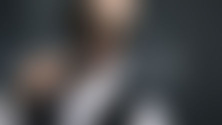 Man in sunglasses blows smoke ring, holding cigarette. Wears white shirt, black vest. Dark background, relaxed and confident mood.