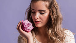 Young woman with long hair gently holding a pink rose, symbolizing balance, beauty, and female hormones, against a soft purple background.
