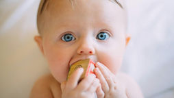 Baby with blue eyes chewing a wooden block. Light background creates a soft and serene mood. Block shows a faint circular design.