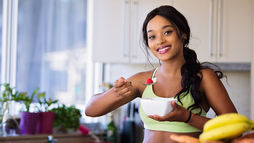 Woman smiling in a bright kitchen while holding a bowl and spooning a strawberry, surrounded by fresh fruits, representing healthy lifestyle habits.