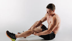 Shirtless man sitting on floor holding his leg in pain, wearing black shorts and orange-soled shoes, against a plain white background.