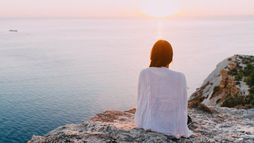 Person in white sitting on a cliff, gazing at a serene ocean sunset, symbolizing reflection and balanced mental wellness.