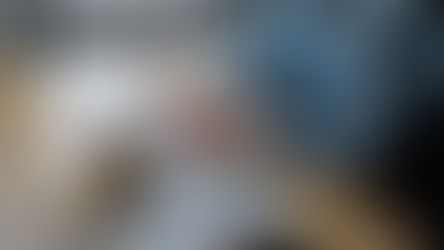 Person in blue shirt typing on a laptop at a wooden desk with notebooks, a cup of coffee, and a window in the background. Calm atmosphere.