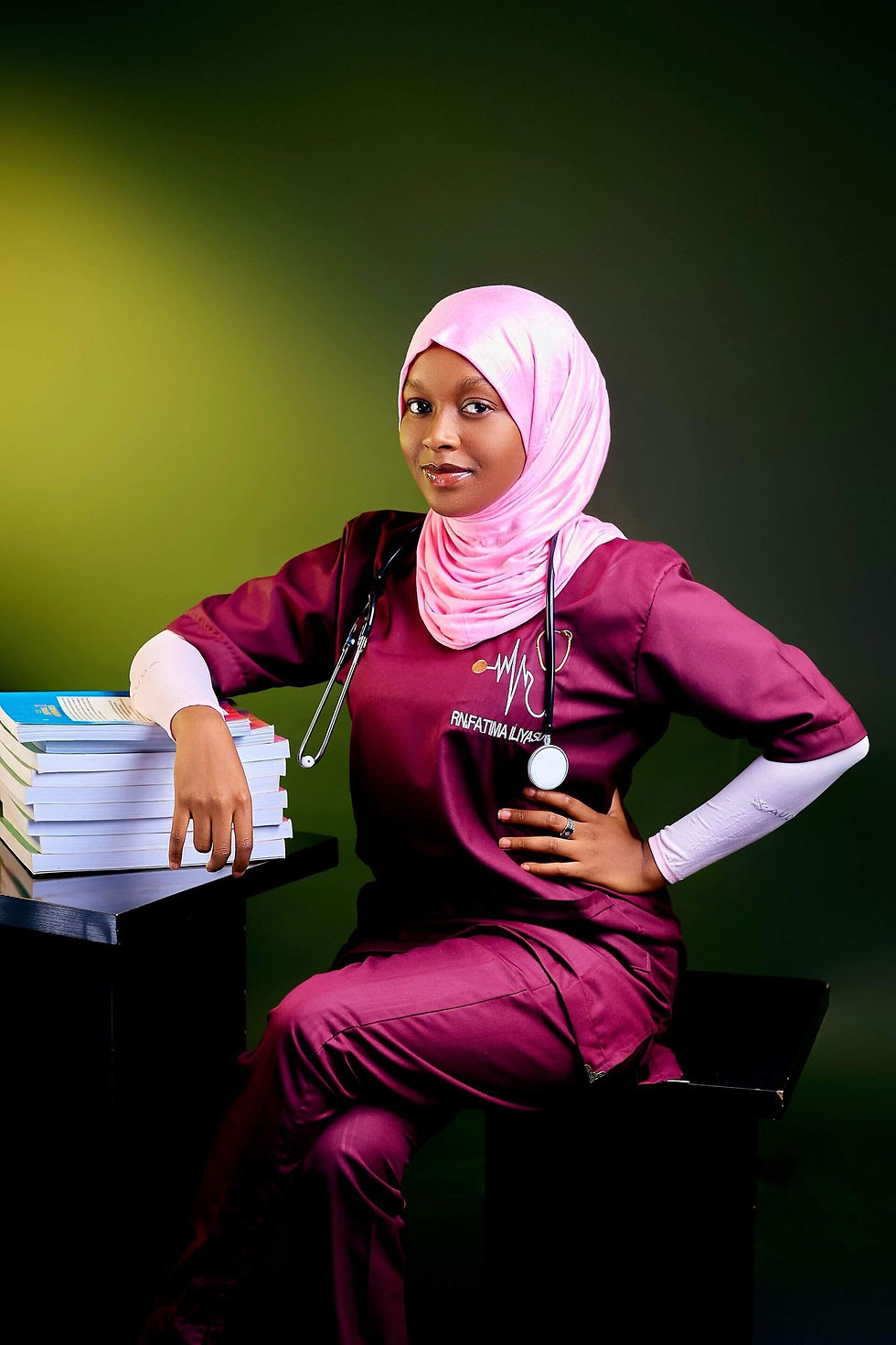 Smiling nurse in pink hijab and maroon scrubs with stethoscope leans on stacked books against a green backdrop.