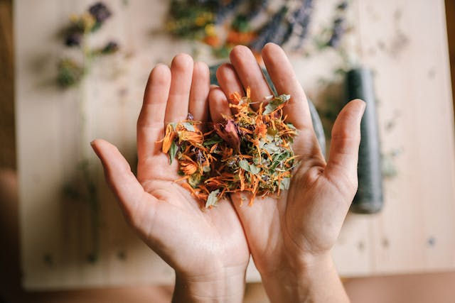 Hands holding colorful dried flowers, on a wooden table with more herbs in background. Natural and calm setting.