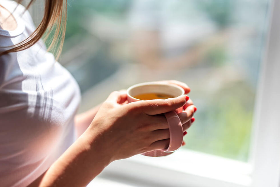 Person in white shirt holding a pink mug with tea by a window. Sunlight streams in, creating a peaceful, reflective mood.