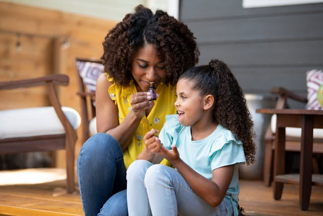 Mother and daughter apply nail polish on a wooden deck, smiling. The mother wears a yellow shirt, daughter in blue. Cozy outdoor setting.