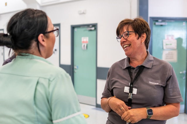 Two women smiling and talking in a hospital corridor. One wears a green uniform, the other a "Volunteer" shirt. Doors with signs in the background.