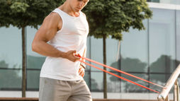 Man exercising with resistance bands outdoors, wearing white tank and gray shorts. He's demonstrating infraspinatus exercises—part of an infraspinatus muscle workout to strengthen shoulder stability. Background of trees and glass building, sunny day.