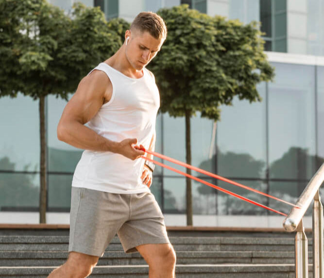 Man exercising with resistance bands outdoors, wearing white tank and gray shorts. He's demonstrating infraspinatus exercises—part of an infraspinatus muscle workout to strengthen shoulder stability. Background of trees and glass building, sunny day.
