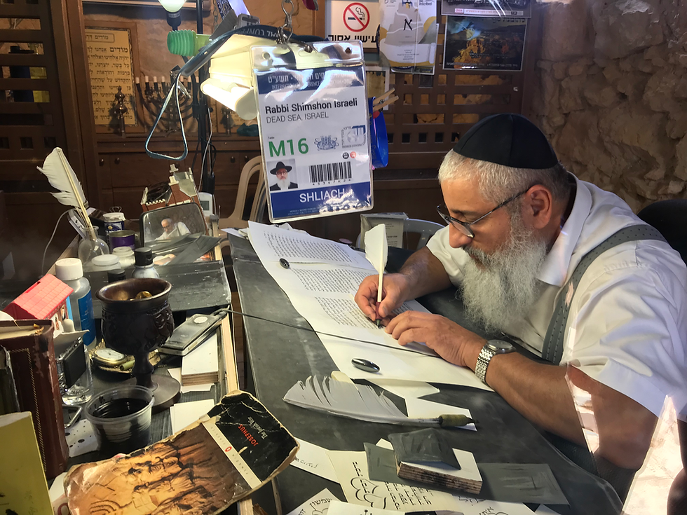 Man with a kippah writing with a quill in a cozy, cluttered room. ID tag hangs nearby. Warm lighting, contemplative atmosphere.