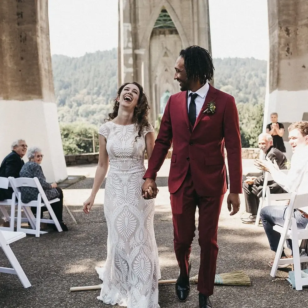 A bride and groom in a vibrant red suit and a white dress stepping over a broom during a wedding ceremony.