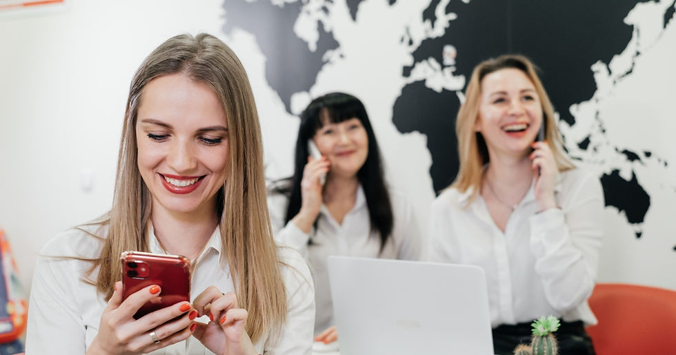 Three women in white shirts smiling, using phones in an office with a black world map on the wall. Red accents and a relaxed mood.