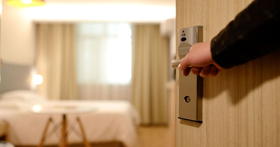 Hand opening hotel room door, revealing a blurred, warmly lit interior with a bed and table. Wooden door contrasts white bedding.
