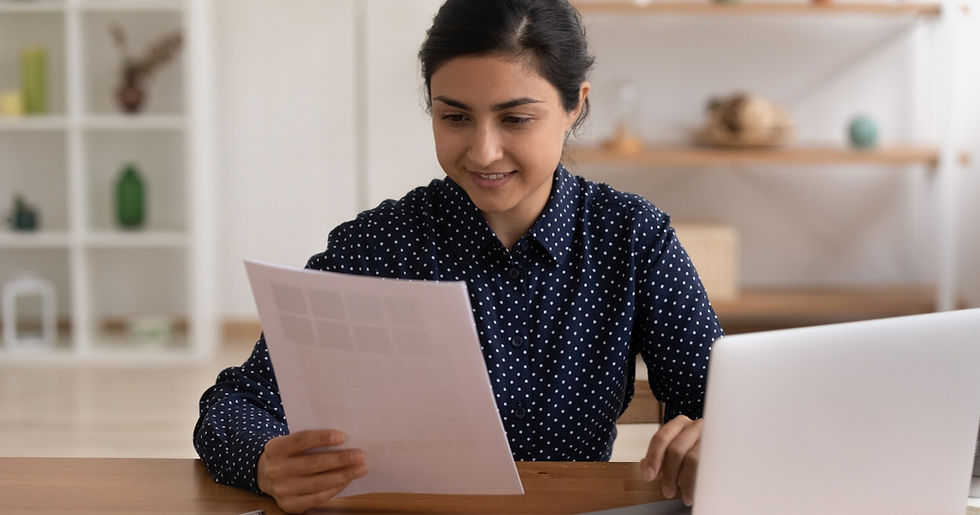 Woman in polka-dot shirt reads a document at a desk with a laptop. Shelves with decor are in the blurred background. She looks focused.