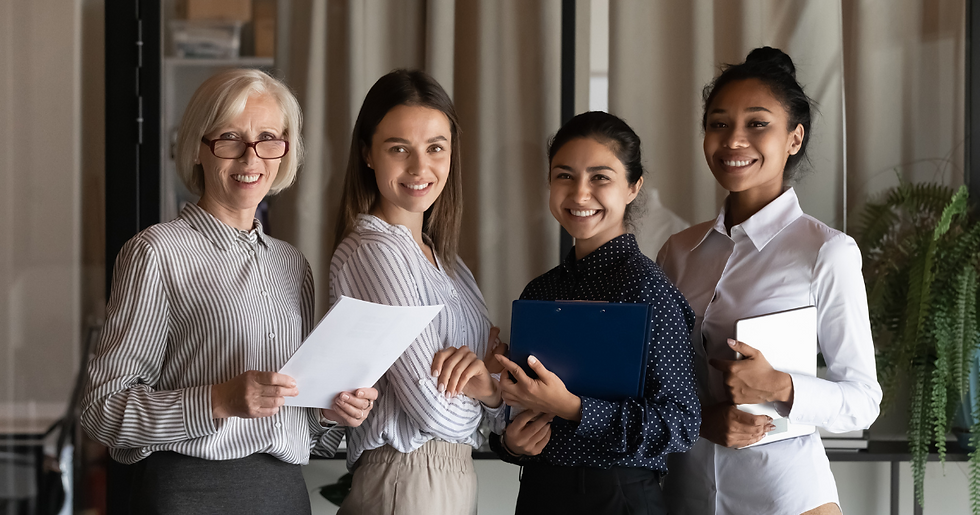 Four women smiling in an office setting, holding documents and a clipboard. They're dressed in business attire, standing near a curtain.