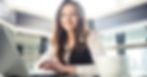 Smiling woman in office attire using a laptop at a bright, modern workspace. Blurred white mug in foreground. Mood is professional and positive.