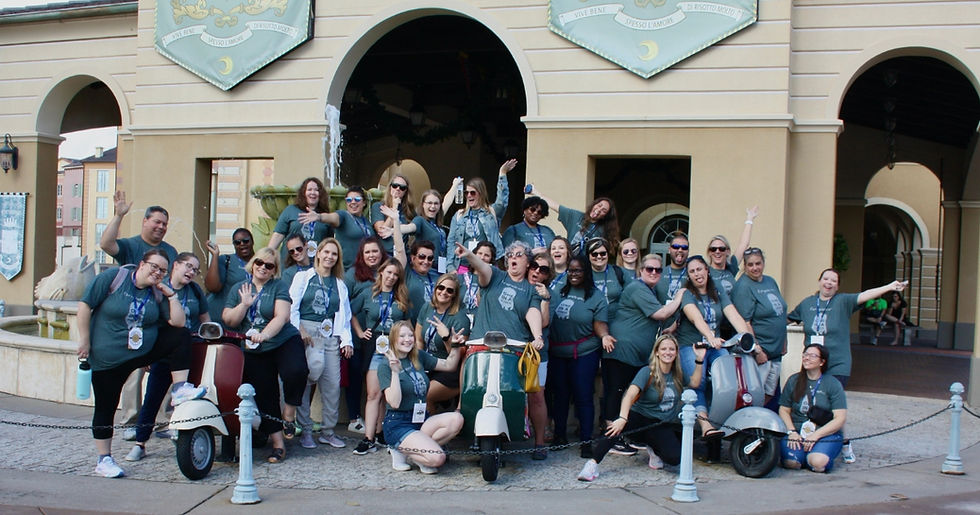 Group of people in gray shirts posing excitedly with scooters in front of an archway with banners. Lively and cheerful atmosphere.