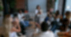 A woman stands smiling, holding a tablet, while a diverse group sits in a circle clapping. Room has brick walls and plants, relaxed vibe.