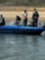 Men fishing on a blue Angler boat on a river, one casting a rod, with a grassy shoreline behind.