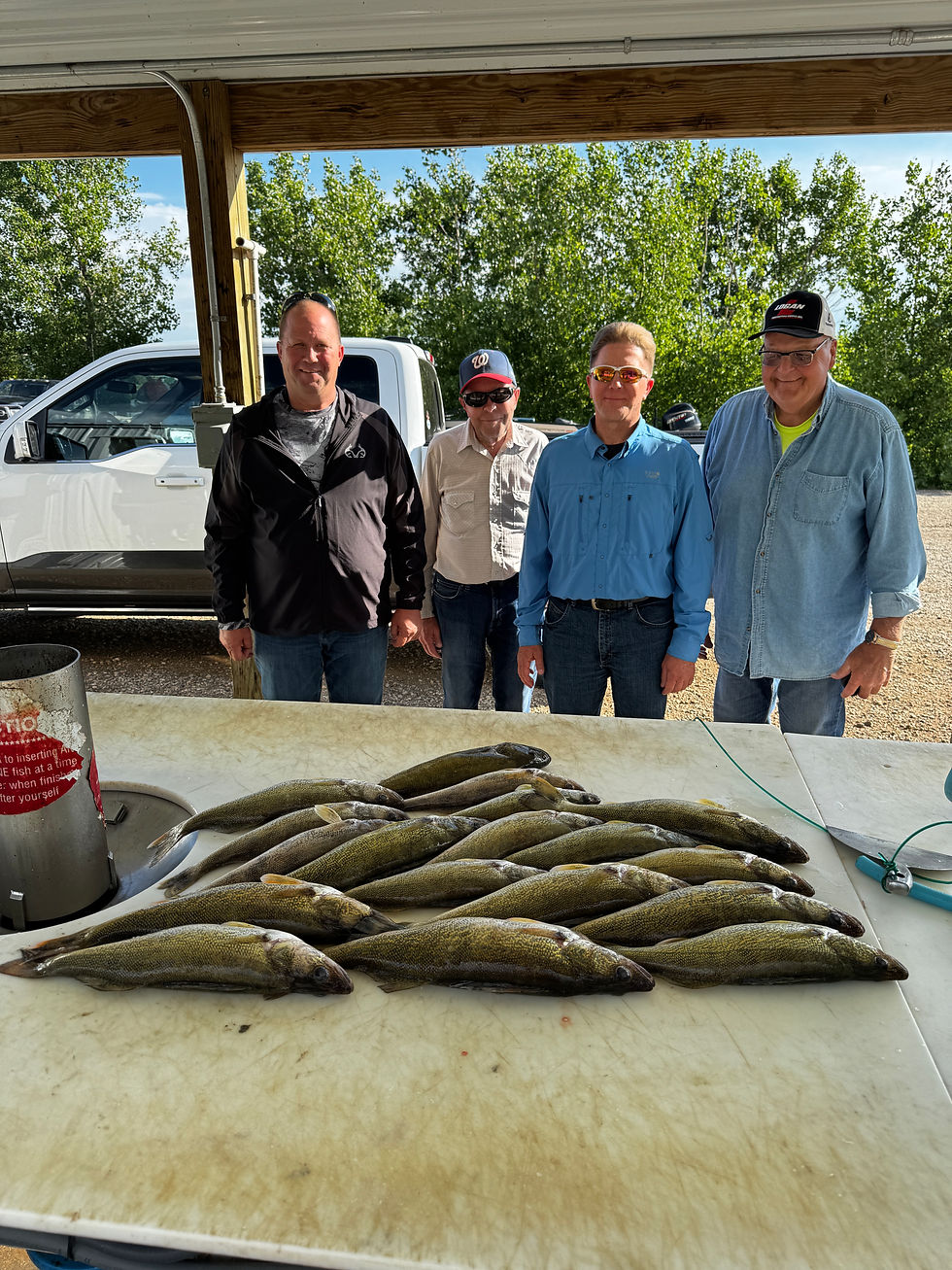 Four men pose behind a table piled with freshly caught fish outdoors, beside a white pickup and trees.