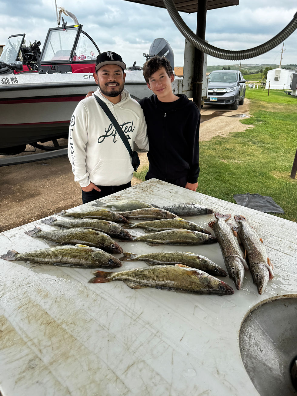 Two smiling people stand by a boat beside a table covered with fresh fish at a lakeside camp, with cloudy sky and parked car.