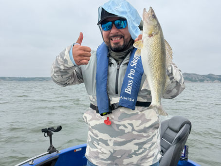 Smiling angler on a boat holds up a fish and gives a thumbs-up, wearing a Bass Pro Shops vest on a cloudy lake sakakawea.