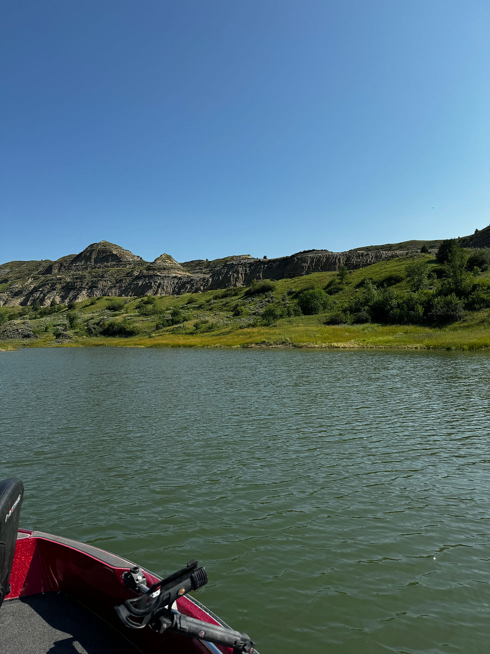 View from a red boat across a calm lake toward green hills and rocky cliffs under a clear blue sky