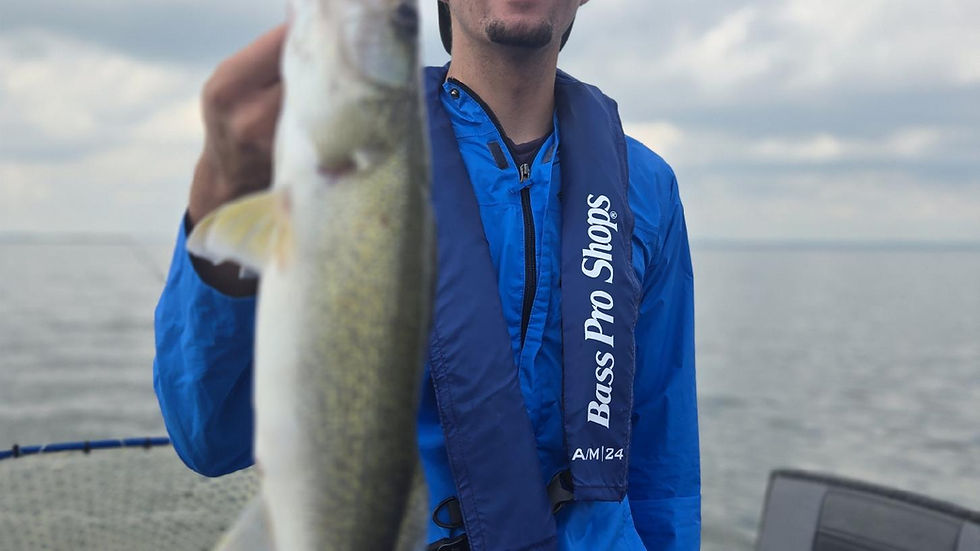 Smiling man on a boat holds up a fish; his blue life vest reads Bass Pro Shops A/M24, with calm water and cloudy sky behind.