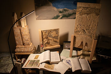 Warm lighting in a dark room that looks like a study with a view of what's on the desk, maps, books, plywood carvings