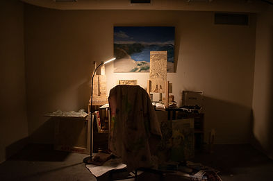 Warm lighting in a dark room that looks like a study shot from behind of the desk and chair, there's a painting on the wall