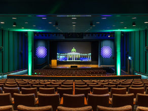 Empty auditorium with rows of seats facing a lit stage and screen showing a domed building, teal and purple lights.
