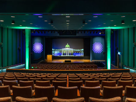 Empty auditorium with rows of seats facing a lit stage and screen showing a domed building, teal and purple lights.