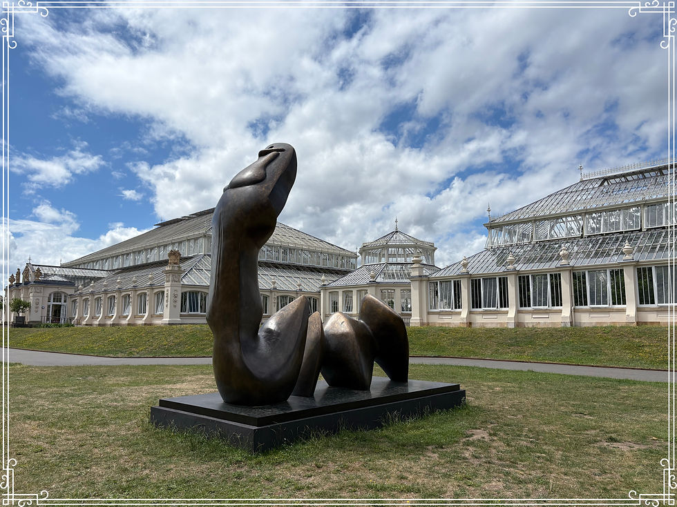 Abstract bronze sculpture by Henry Moore on a lawn in front of glass conservatories under a cloudy blue sky, with ornate white border framing the image