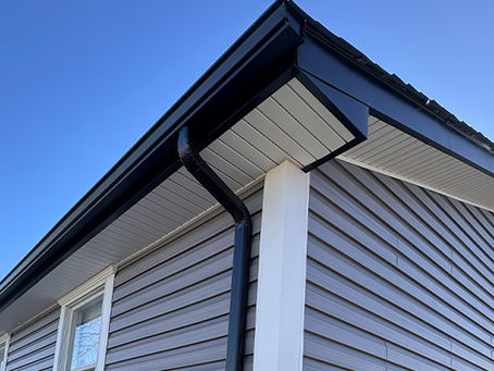 A black rain gutter and downspout running along the edge of a house roof, attached beneath the eaves of a gray-sided home against a clear blue sky.