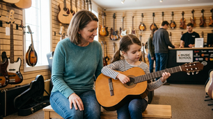 A young girl strums a classical guitar in a music store next to her encouraging mother, surrounded by instruments on display and a store counter in the background.