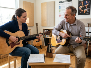 A focused adult student and an encouraging music teacher sitting together in a bright, modern practice room during an engaging private lesson. The student holds an acoustic guitar, carefully watching the teacher demonstrate a fingering technique on another guitar. On a small table nearby sits an open notebook with written questions, a metronome, and a music score, highlighting a structured and interactive learning environment.